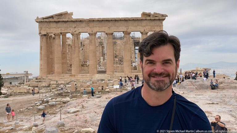 Smiling male visitor in front of the Parthenon, with other tourists and rocky Acropolis ground behind.