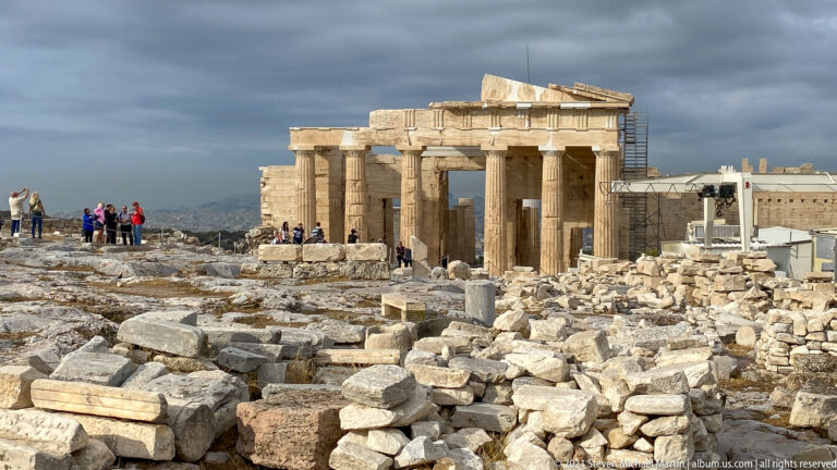 Propylaia rising beyond scattered marble blocks and foundations, with visitors gathered near the gateway.