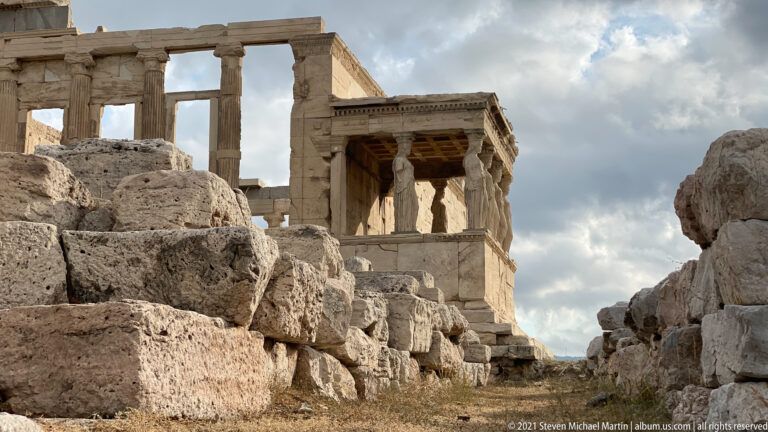 Caryatid copies on the Erechtheion’s south porch glimpsed through rough stone walls and rocky Acropolis ruins.