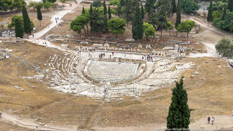Elevated view of the Theatre of Dionysus with semicircular stone seating, orchestra area, ruins, and trees.