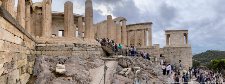 Visitors climbing stone steps through the Propylaia, with tall columns and the Temple of Athena Nike beyond.