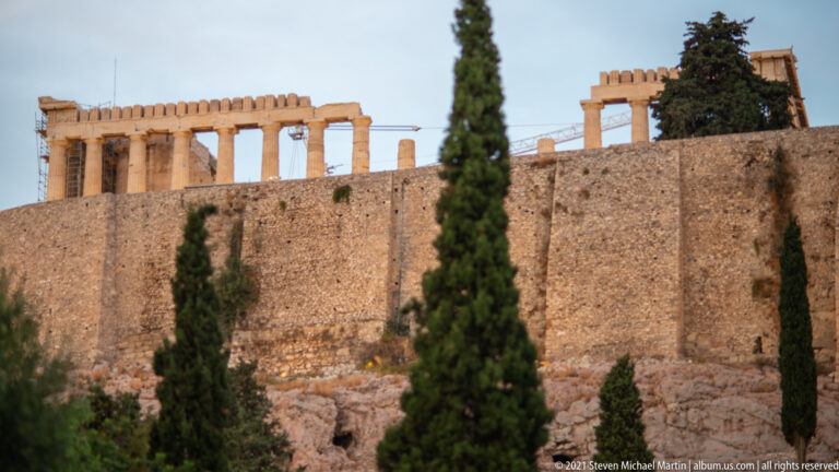 Parthenon columns visible above a tall Acropolis stone wall, with cypress trees in the foreground.