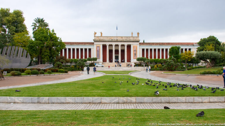 Neoclassical façade of the Hellenic National Archaeological Museum in Athens behind a landscaped forecourt and central columned portico.