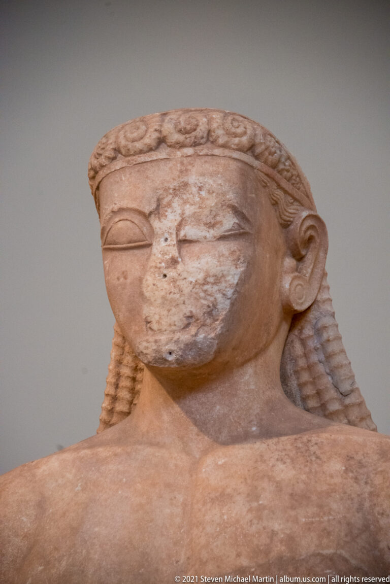 Close view of an Archaic male sculpted head with long patterned hair and almond-shaped eyes.