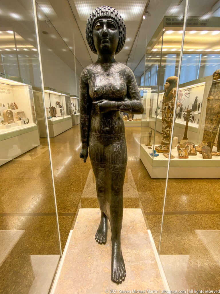 Standing bronze female statuette with patterned hair, long fitted garment, one arm across the torso, and one arm lowered.