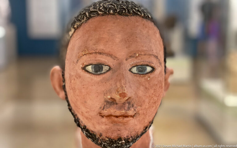 Painted male head with large outlined eyes, short curly hair, reddish skin tones, and a narrow beard along the jaw.