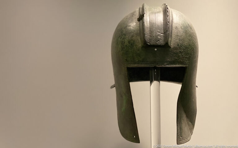 Green-patinated bronze helmet with rounded crown, long cheek pieces, and a raised vertical strip over the top.