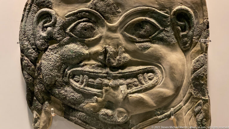 Bronze relief face with bulging eyes, broad nose, wide tooth-filled mouth, and heavy green-black patina.