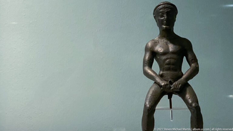 Small bronze nude male figure seated frontally with knees apart and both hands lowered toward the groin.