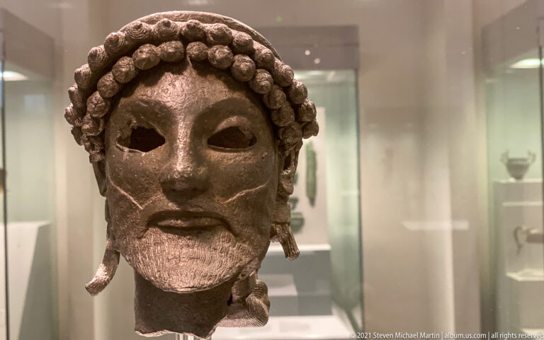 Bronze bearded male mask with hollow eyes, rounded curls, modeled beard, and projecting side elements.