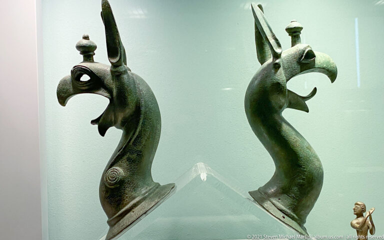 Pair of bronze griffin heads with long curved necks, hooked beaks, pointed ears, open jaws, and green patina.