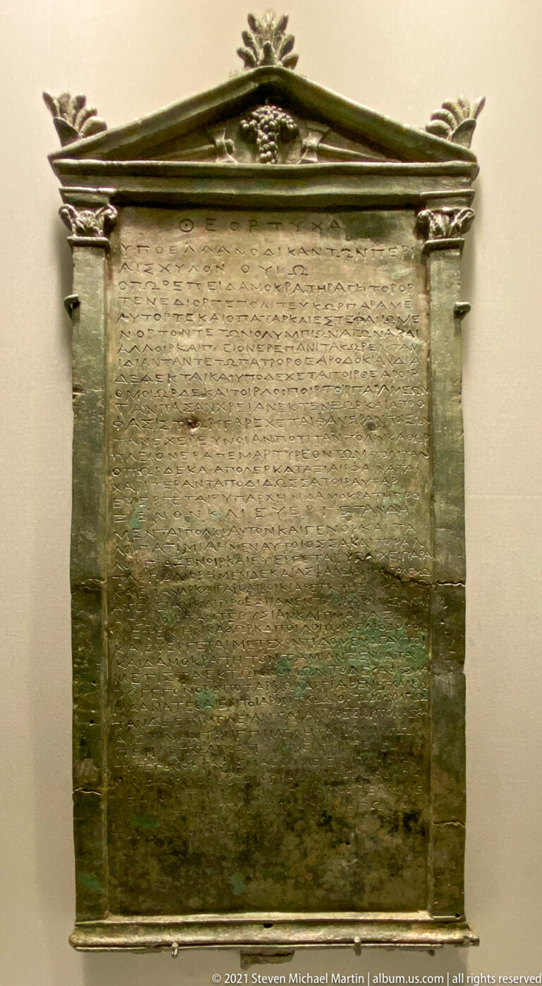 Rectangular bronze tablet with many lines of Greek inscription, framed by molded sides and a pediment-like top.