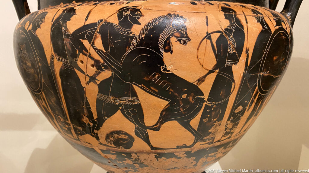 Black-figure vase scene of a muscular hero wrestling a lion, with additional armed and draped figures nearby.