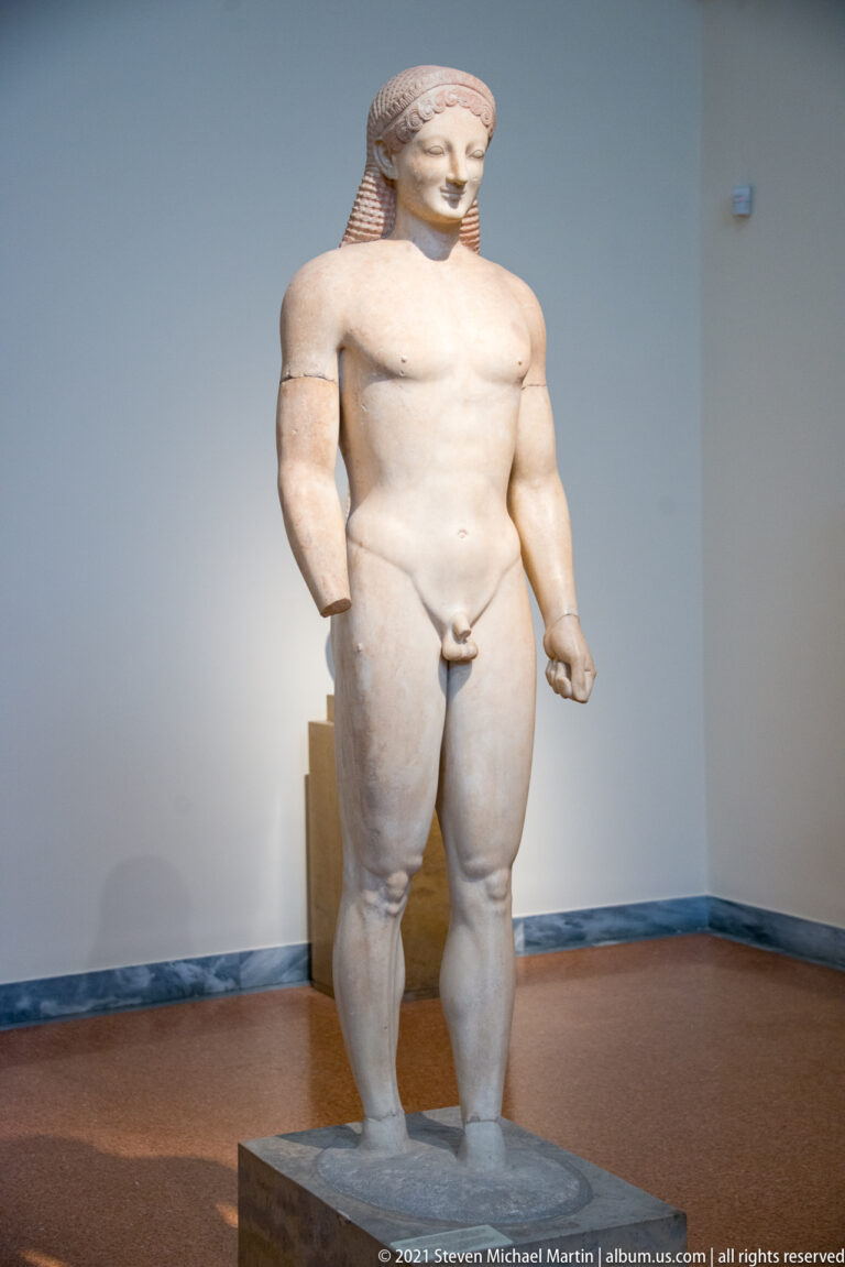 Nearly complete nude kouros standing frontally with one leg advanced and long patterned hair.