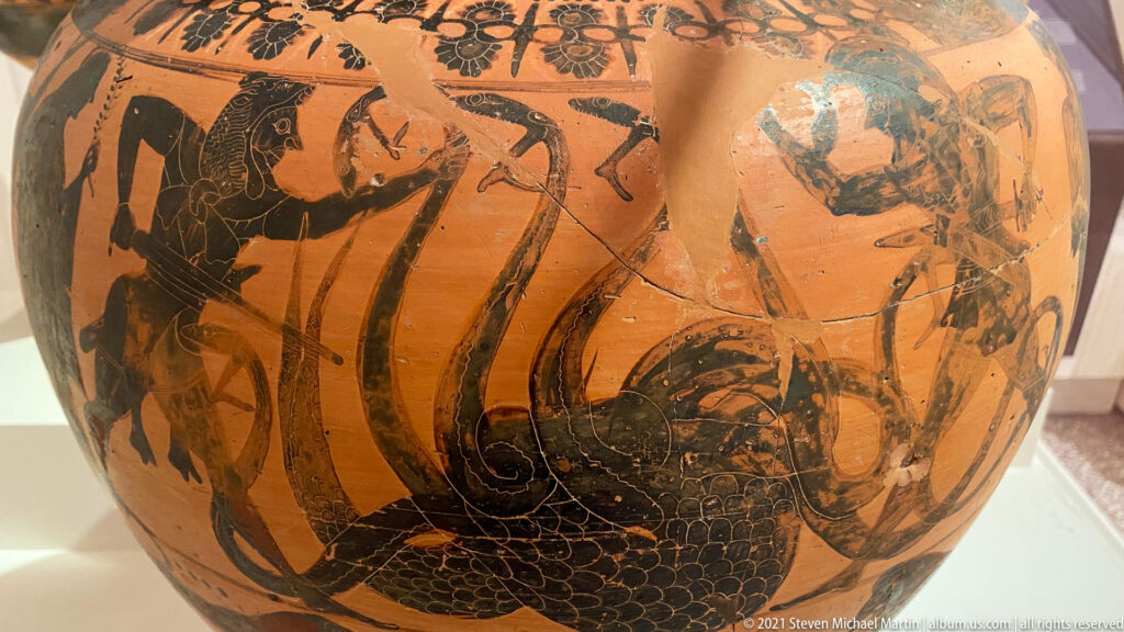 Black-figure vase with a large coiling scaled serpent or sea creature, chariot elements, and surrounding human figures.