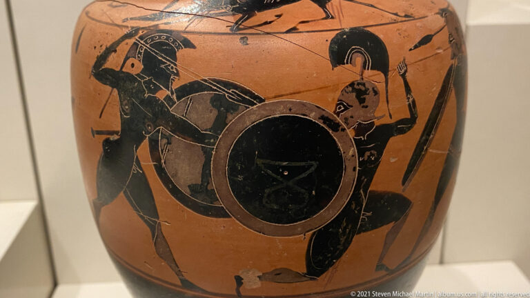 Black-figure vase scene of two armored warriors fighting with large round shields, helmets, and spears.