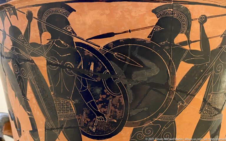 Black-figure vase detail of a helmeted warrior with a large round shield surrounded by other armed figures.