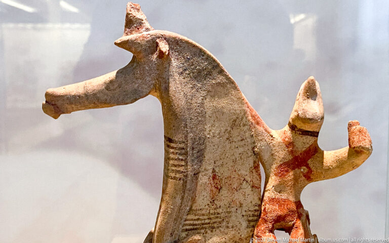 Painted terracotta horse with long muzzle and pointed ears beside a small human-like figure with raised arms.