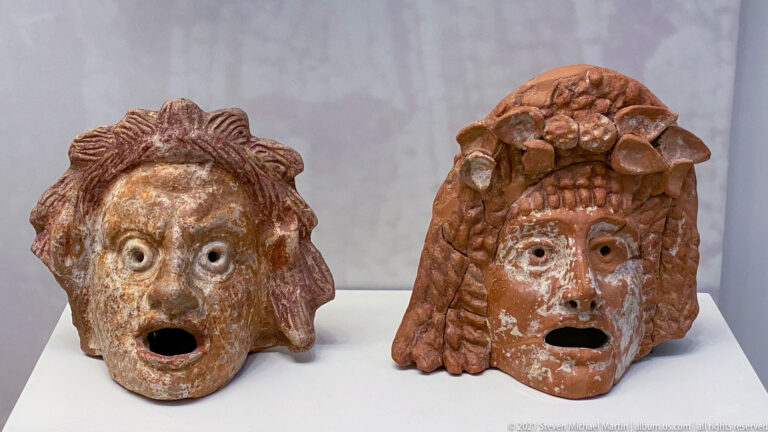 Two terracotta masks with wide open mouths, large eyes, and different modeled hairstyles displayed side by side.
