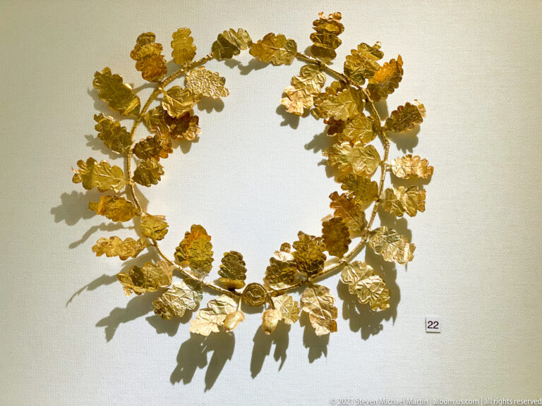 Gold wreath made from many thin oak-like leaves and small acorn-like fruits arranged around a circular stem.