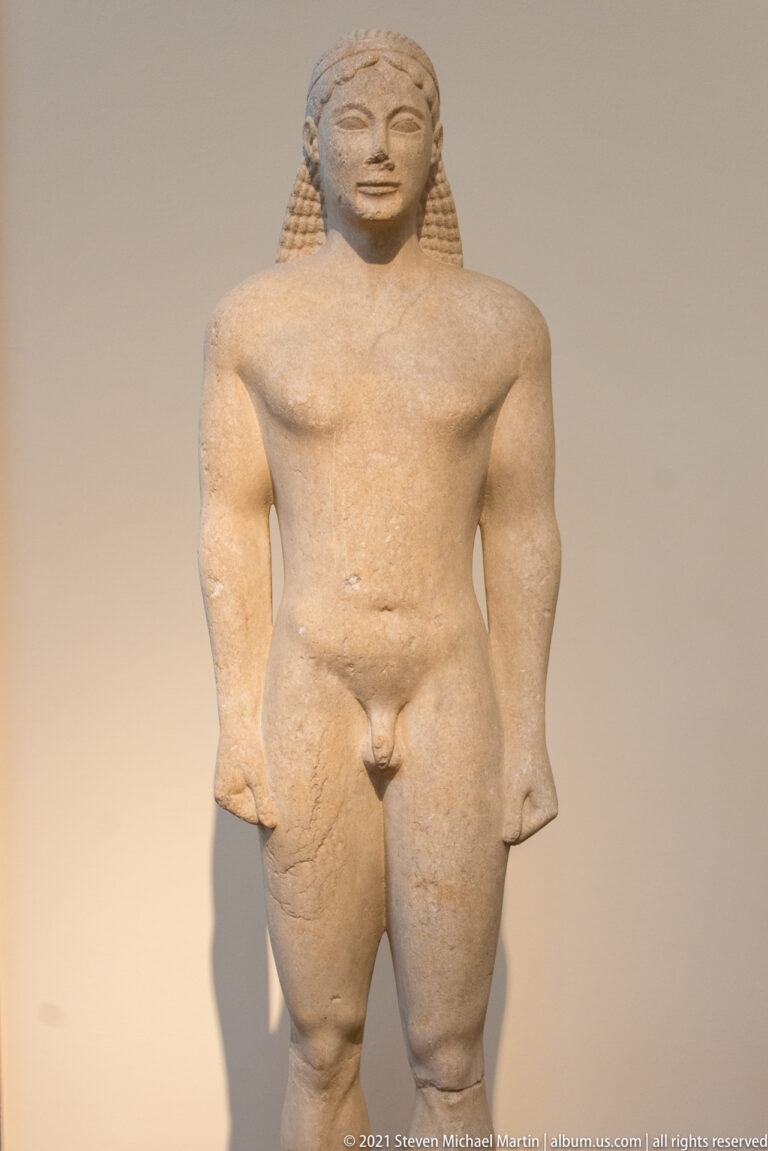 Stocky nude kouros standing frontally with thick limbs, patterned hair, and arms at sides.