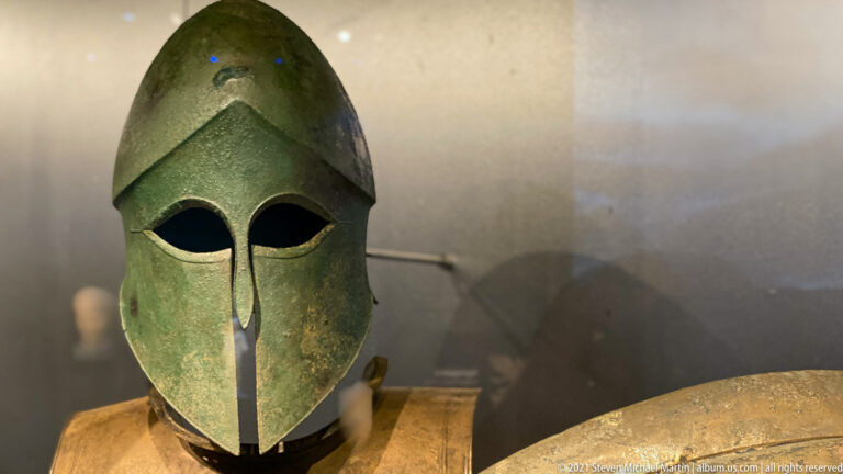Green bronze Corinthian-type helmet with almond-shaped eye openings, smooth crown, and long central nose guard.