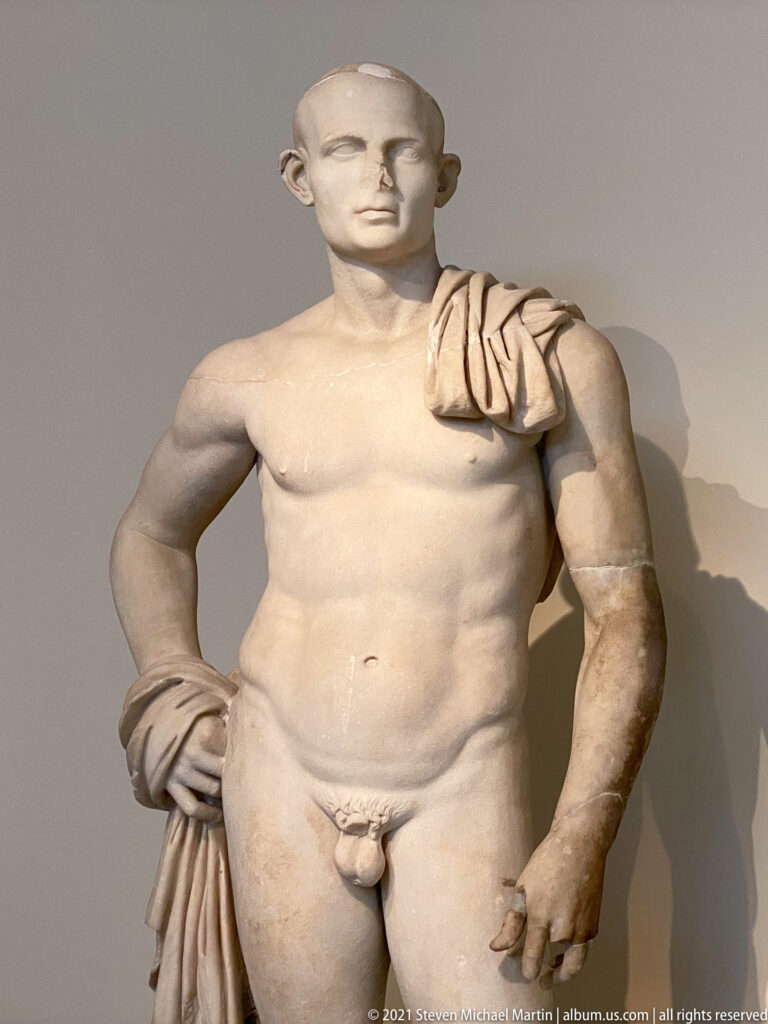 Stocky marble nude male statue with short hair, drapery over one shoulder, and one hand resting at the hip.