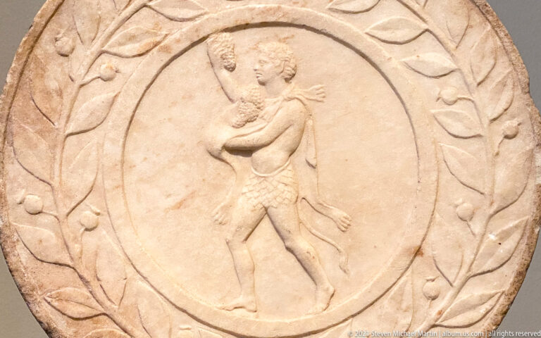 Round marble relief with a standing youthful male carrying a long object, surrounded by a carved leafy border.