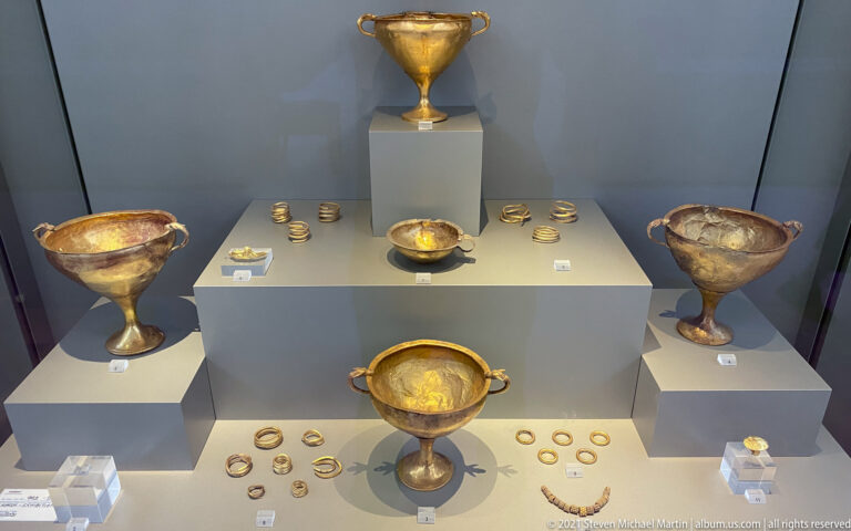 Display case of Mycenaean gold stemmed cups, handled bowls, and small gold ornaments arranged on gray plinths.