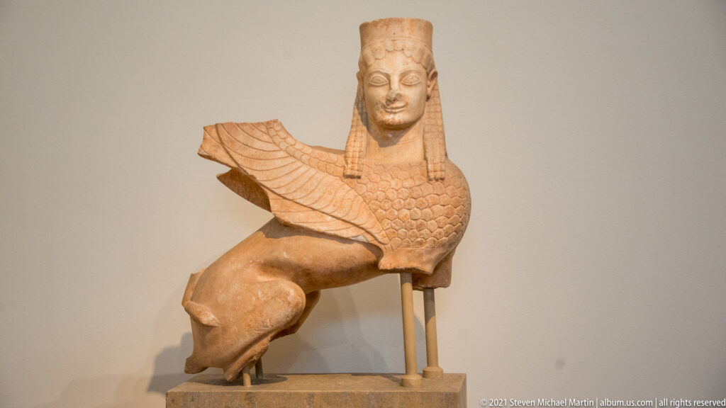 Archaic stone sphinx with female head, wing, and seated feline body.