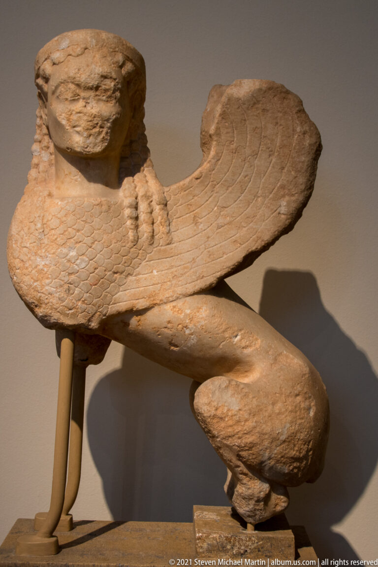 Side view of a stone sphinx showing broad wing, human head, and crouched feline body.