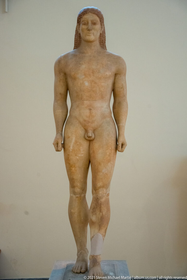Front-facing kouros with clenched hands, broad chest, patterned hair, and damaged lower leg.