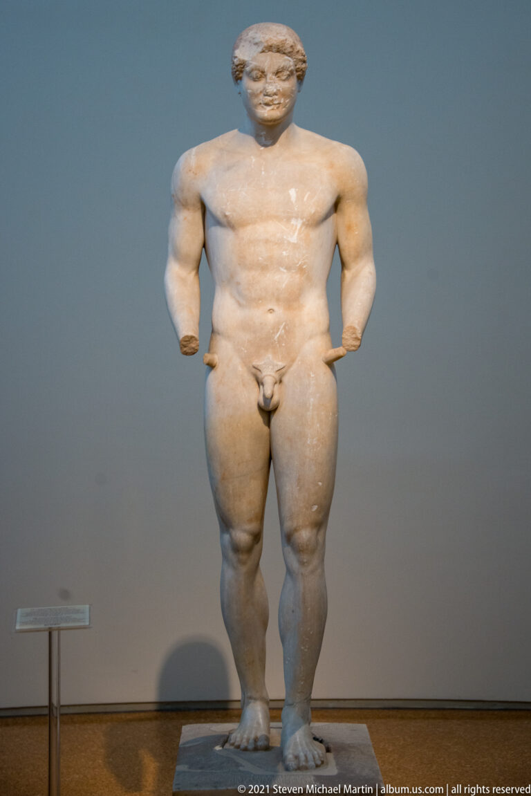 Standing nude male stone statue with short curled hair, muscular torso, and thick legs.