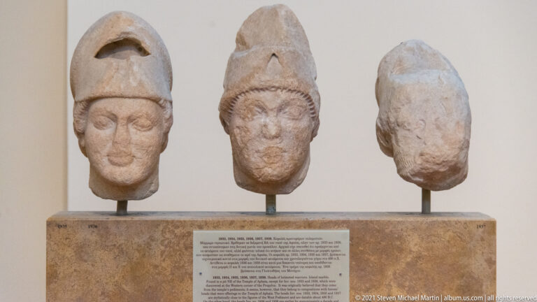 Three ancient stone heads on stands, two wearing tall cap-like headgear.
