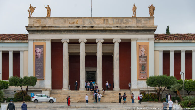 Main entrance portico of the Hellenic National Archaeological Museum with pale columns, broad steps, red wall panels, banners, and visitors.