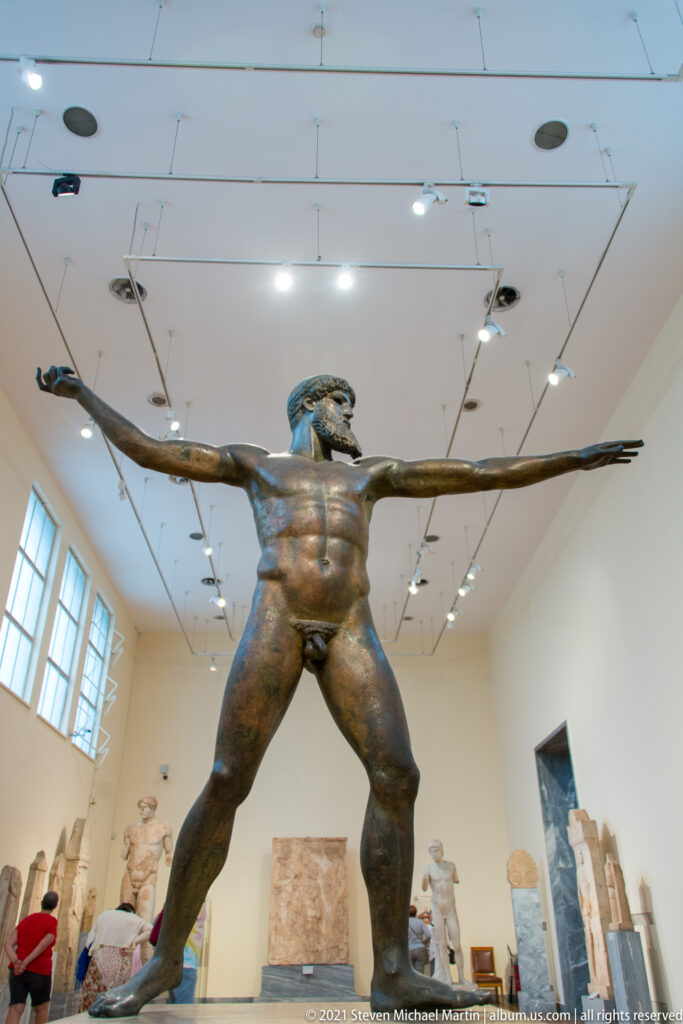 Full frontal view of Zeus or Poseidon of Artemision, a monumental nude bronze with legs spread and both arms fully extended.