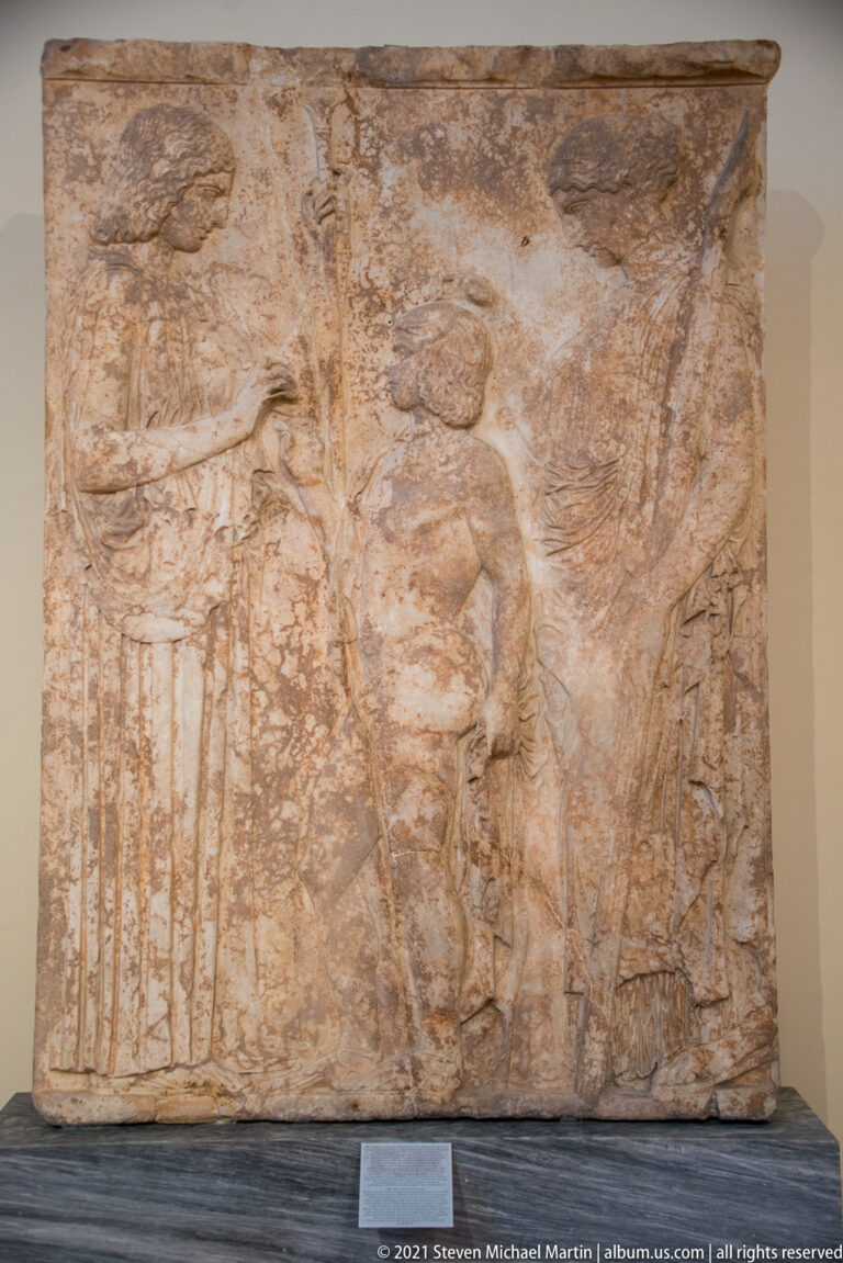 Weathered rectangular stone relief with a standing central figure and faint surrounding carved forms.