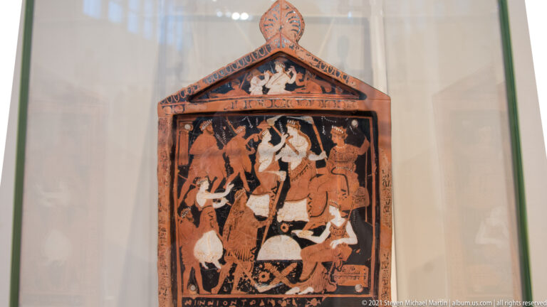 Painted ancient Greek plaque with triangular pediment and crowded mythological figure scene.