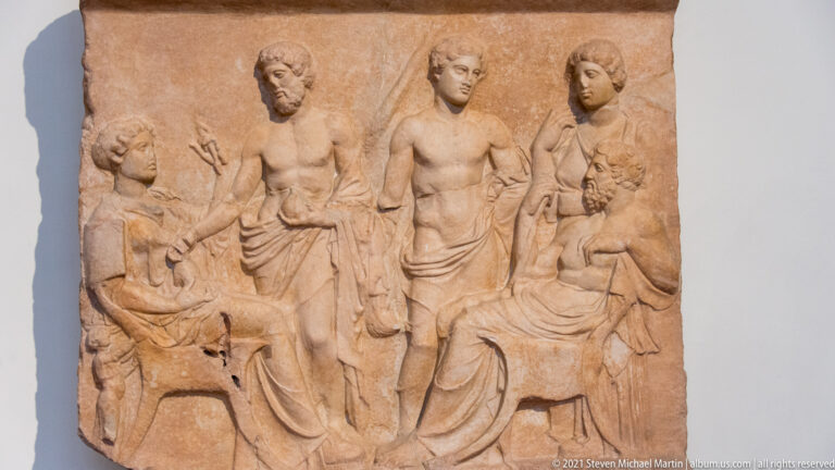 Marble relief with seated and standing adults and children arranged in a multi-figure scene.