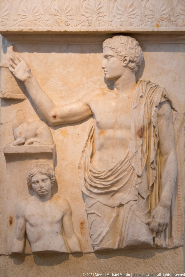 Relief showing a standing draped youth in profile above a smaller figure and carved object.