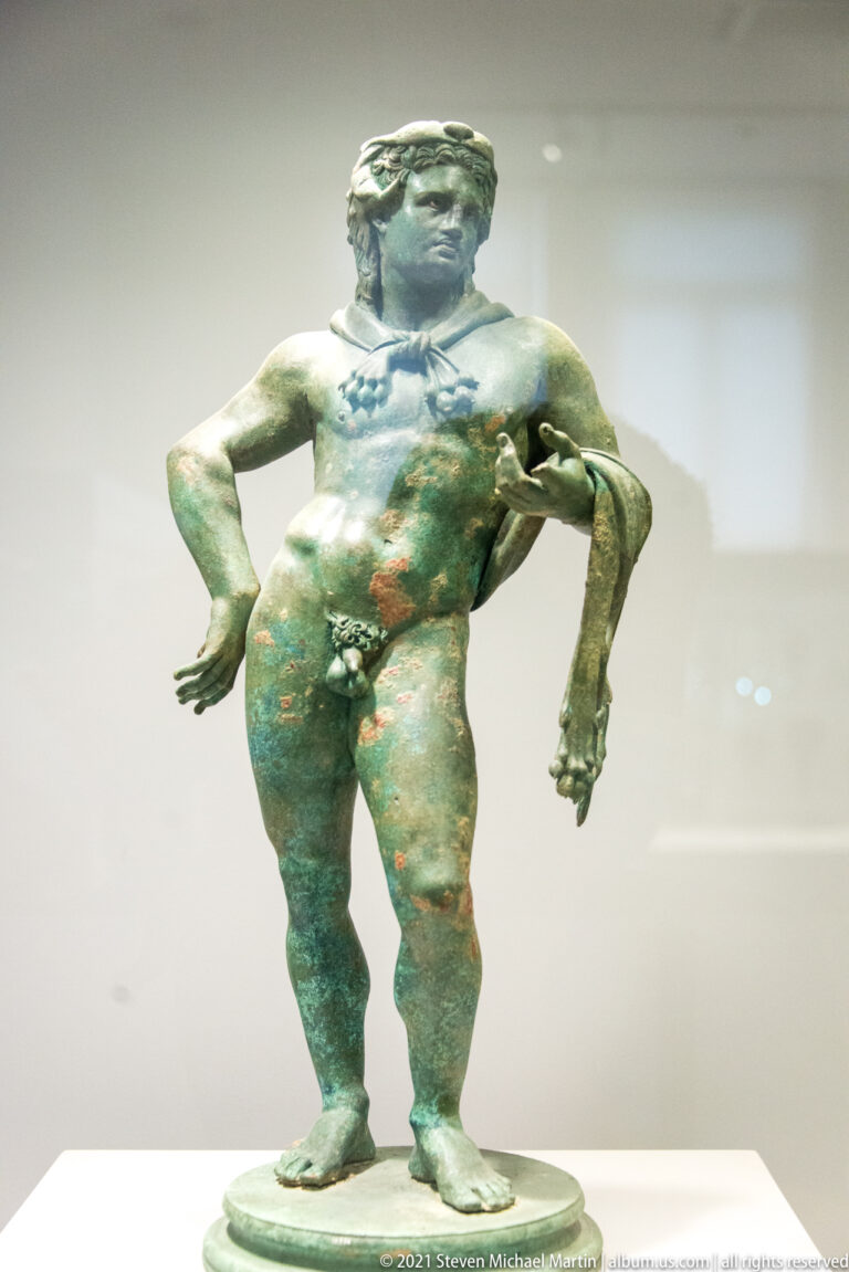Small nude bronze male figure with rounded body, relaxed pose, and green patina.