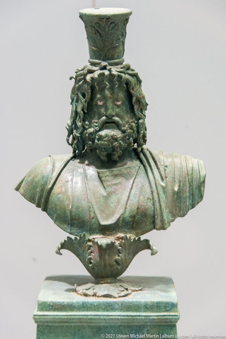 Elaborate green-patinated bronze object with central human bust, floral ornament, and broad projecting elements.