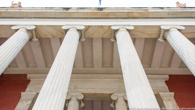 Upward view beneath the Hellenic National Archaeological Museum portico showing fluted columns, capitals, coffered ceiling, and entablature.