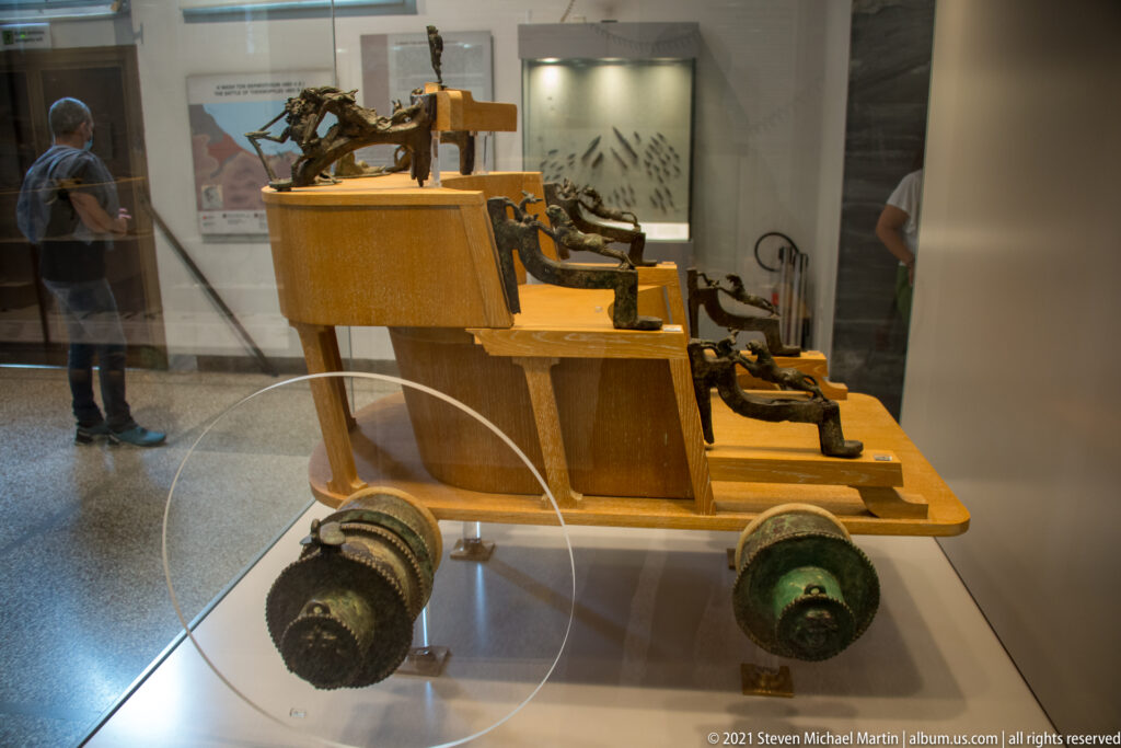 Bronze wheeled chariot model with four wheels, miniature horses, figures, and fittings in a display case.