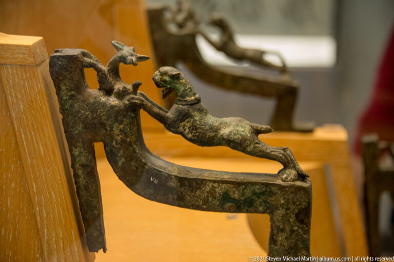 Bronze fitting with reclining nude figure and smaller standing figure on a curved support, displayed in a museum case.