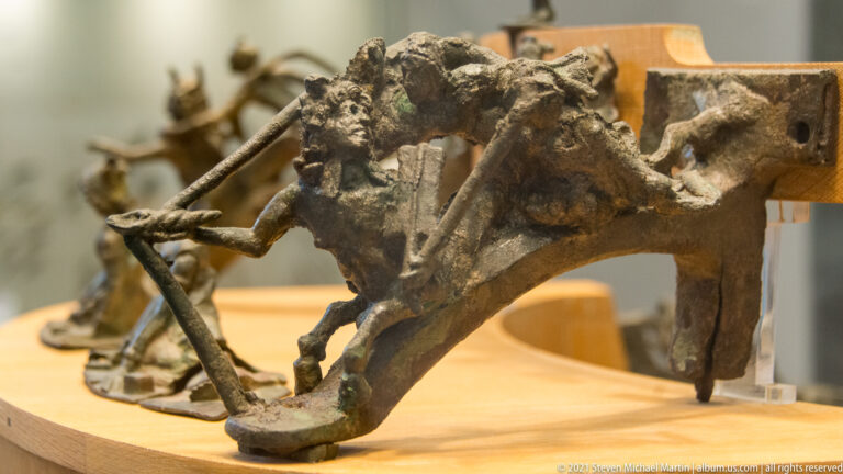 Corroded bronze assembly with rods and plate-like elements mounted on a museum support.