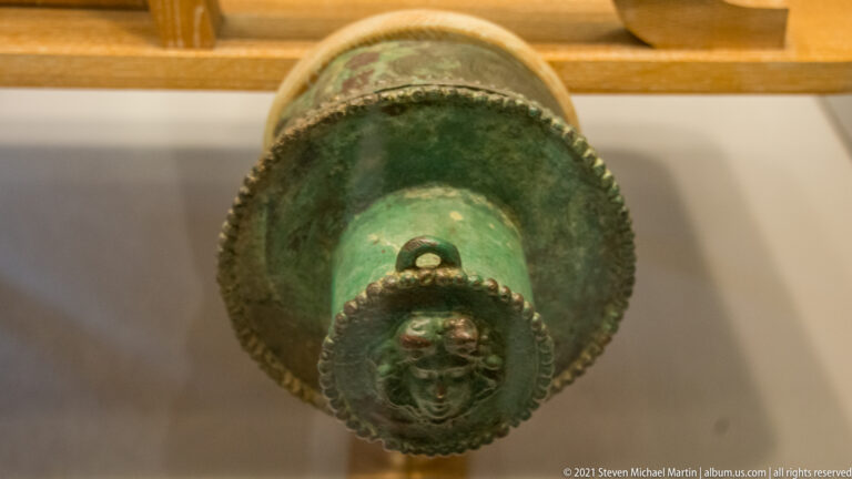 Circular green-patinated bronze element with beaded rim and a projecting face at the center.