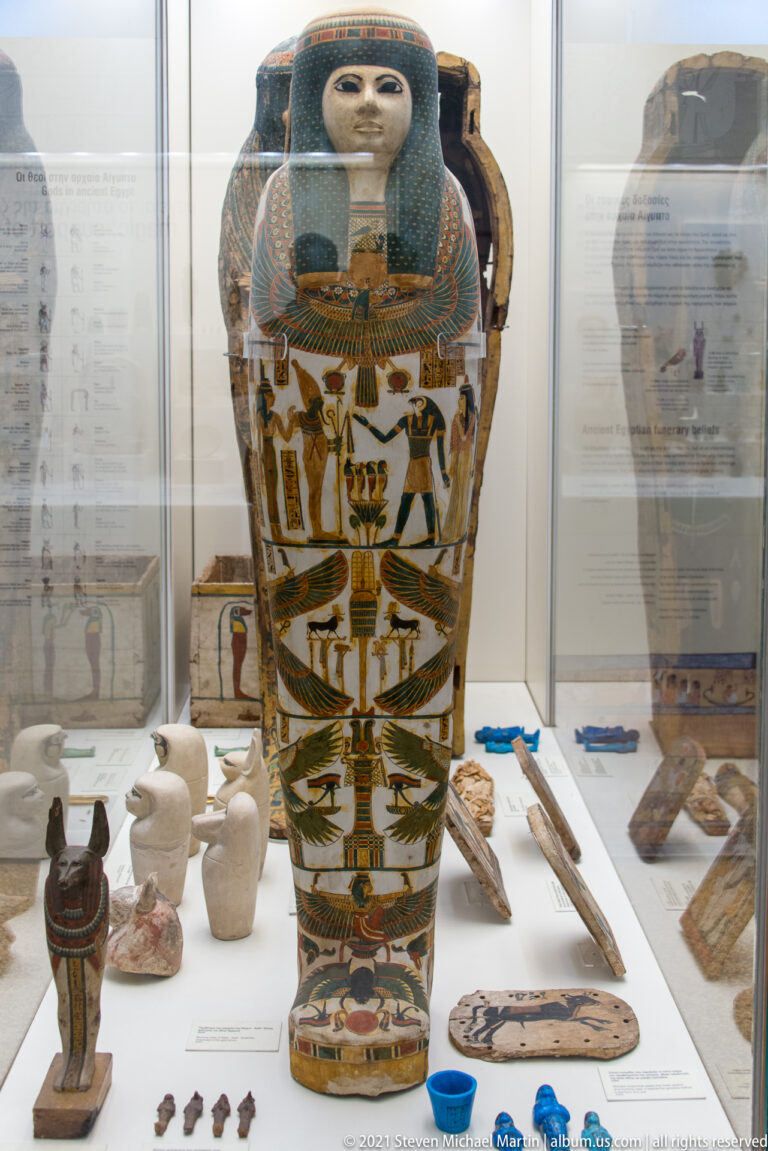 Painted Egyptian anthropoid coffin surrounded by small funerary objects in a glass museum case.