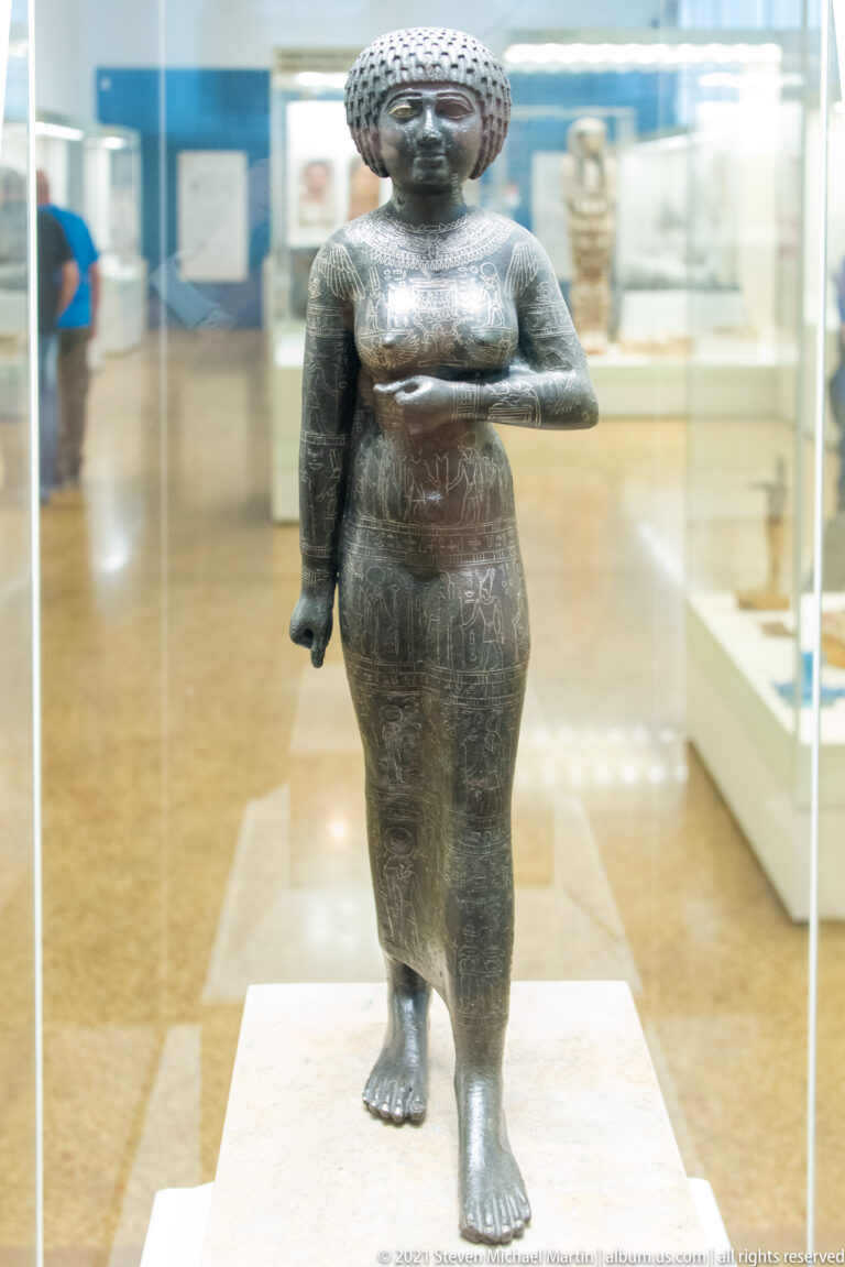 Standing bronze female statuette wearing a patterned headdress and long garment, one arm bent across the torso.