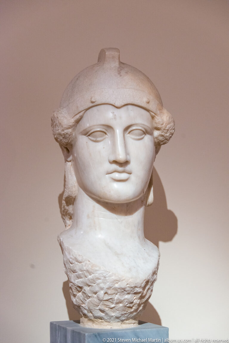 Marble female head wearing a close-fitting helmet, with smooth idealized features and textured carving below the neck.